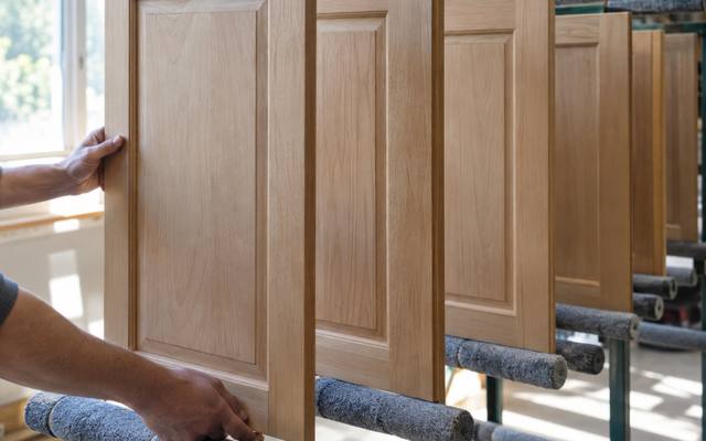 A hand lifting a bare wood cabinet door from a drying rack.