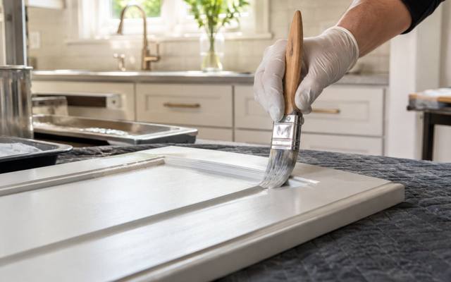 A gloved hand brushing white paint along the edge of a cabinet door on a padded table.